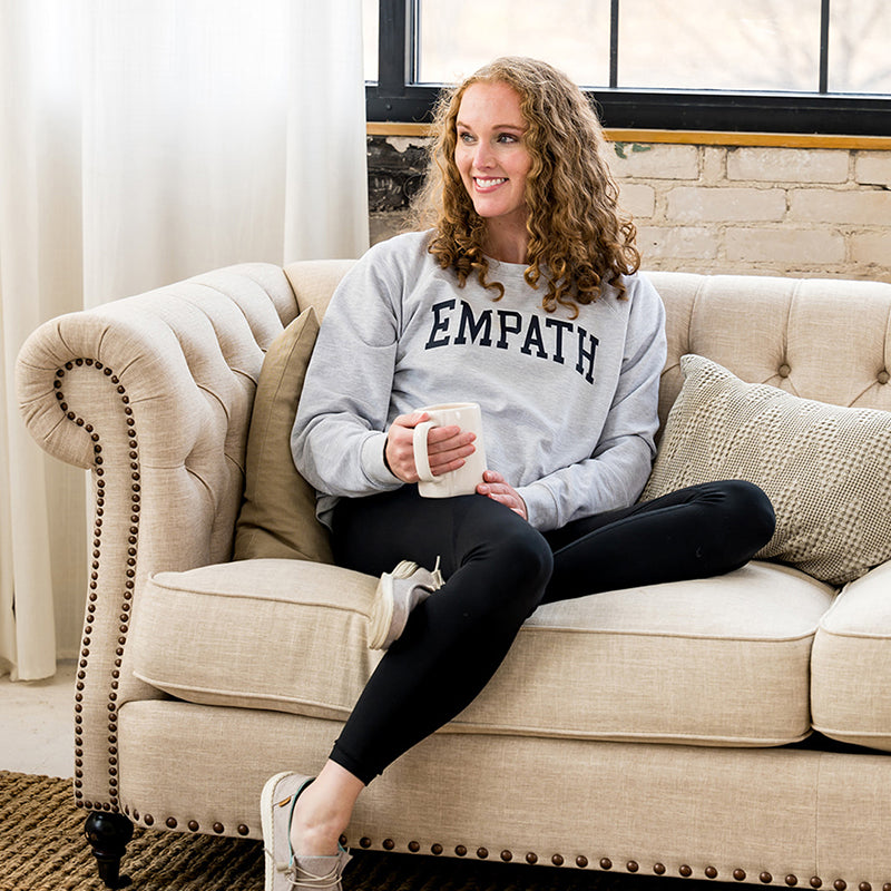 Woman smiling in gray EMPATH crewneck sweatshirt sitting on fabric couch.