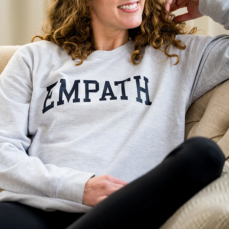 Woman smiling in gray EMPATH crewneck sweatshirt sitting on fabric couch.