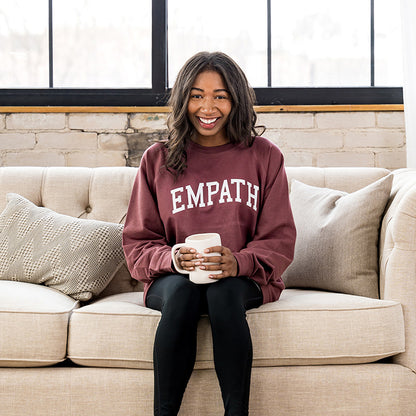 Woman smiling in maroon EMPATH crewneck sweatshirt sitting on fabric couch.