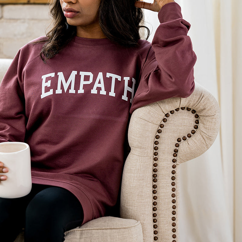 Woman smiling in maroon EMPATH crewneck sweatshirt sitting on fabric couch.