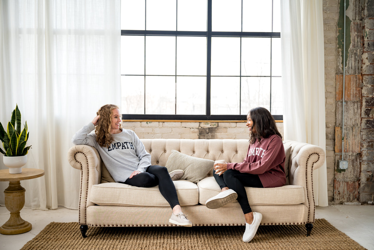 Two women sitting on a fabric couch in an industrial room wearing EMPATH crewneck sweatshirts.