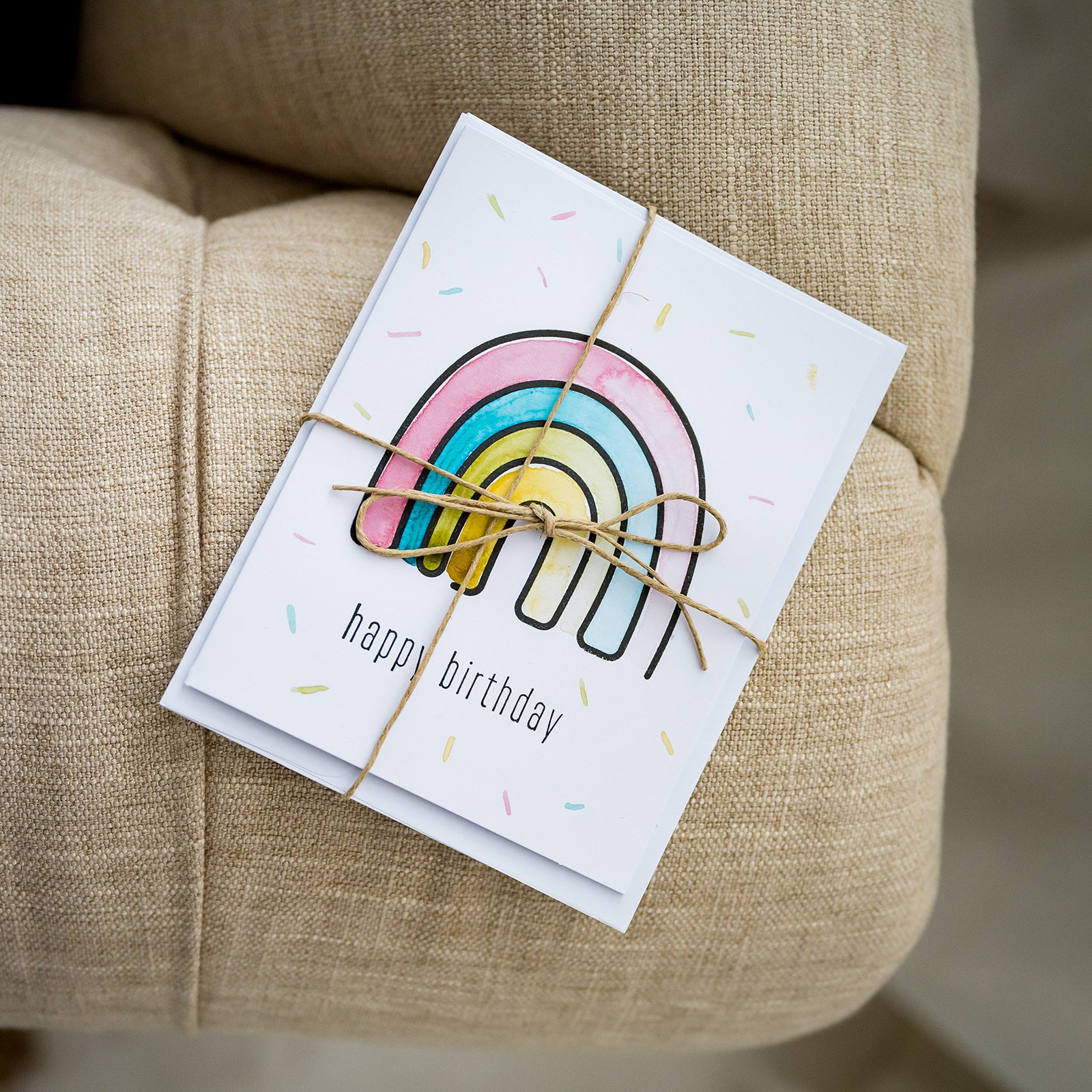 Watercolor and stamped note card of rainbow wrapped in twine on fabric couch.