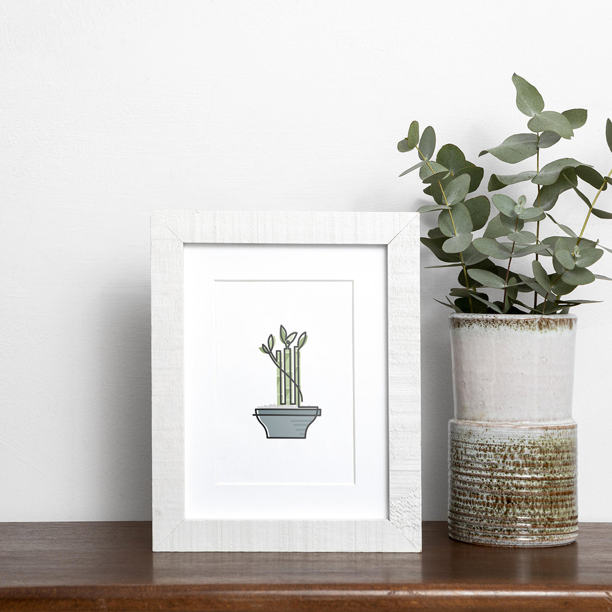 Black single-line art of bamboo in blue pot on white, framed in wood, displayed near vase of eucalyptus on wooden shelf.