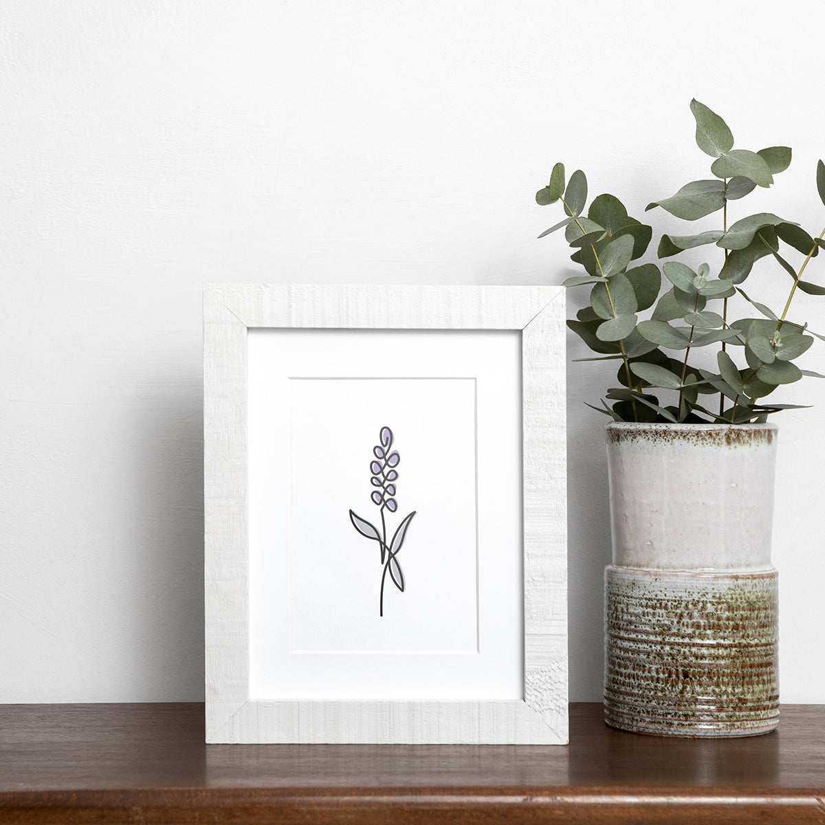 Black single-line art of purple and gray lavender on white, framed in wood, displayed near vase of eucalyptus on wooden shelf.