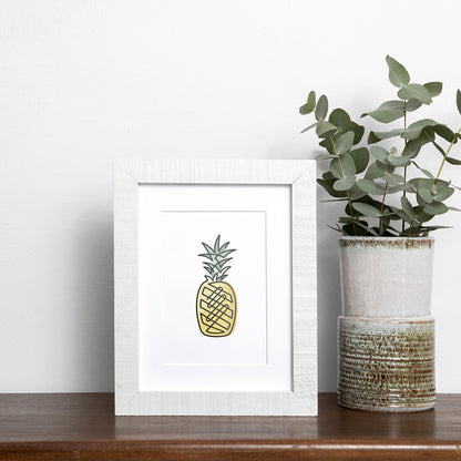 Black single-line art of yellow and green pineapple on white, framed in wood, displayed near vase of eucalyptus on wooden shelf.